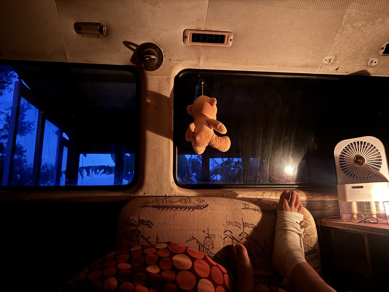 Een beertje hangt aan het raam van een camper — warm kaarslicht binnen, avondblauw buiten